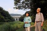 　「中池見湿地」を歩く笹木智恵子さん（左）と進さん。奥は３月に延伸開業した北陸新幹線の高架＝２０２４年７月２３日、福井県敦賀市