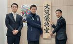 　「大阪儀典室」の看板を持つ、儀典長に任命された植田浩氏（右）と大阪府の吉村洋文知事。左は横山英幸大阪市長＝２０日午前、大阪市住之江区