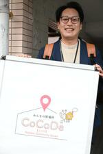 　自身がデザインした「居場所サロン」の看板に手を添えるピアサポーターの寺尾真次さん＝２０２５年３月、鹿児島市