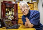　自宅の「定位置」に座る藤野高明。ここでパソコン作業をする。電化製品の場所は固定化し、見えなくても過ごせるよう工夫している。「物の位置は体で覚えています」。仏壇には妻カヨコの位牌（いはい）がある＝２０２５年１月、大阪市（撮影・藤井保政）