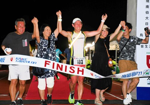  家族らとフィニッシュテープを切る選手（中央）＝１６日午後９時ごろ、米子市のどらドラパーク米子陸上競技場 