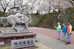 　福井城を築いた結城秀康の像の前で、観光客に説明する語り部（左）＝２０２５年４月、福井市